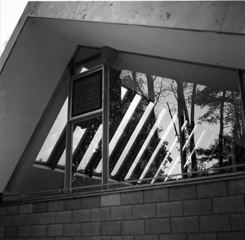 Reflection of trees and outdoor elements in a large trapezoidal window with blinds, on a building exterior with brick wall.