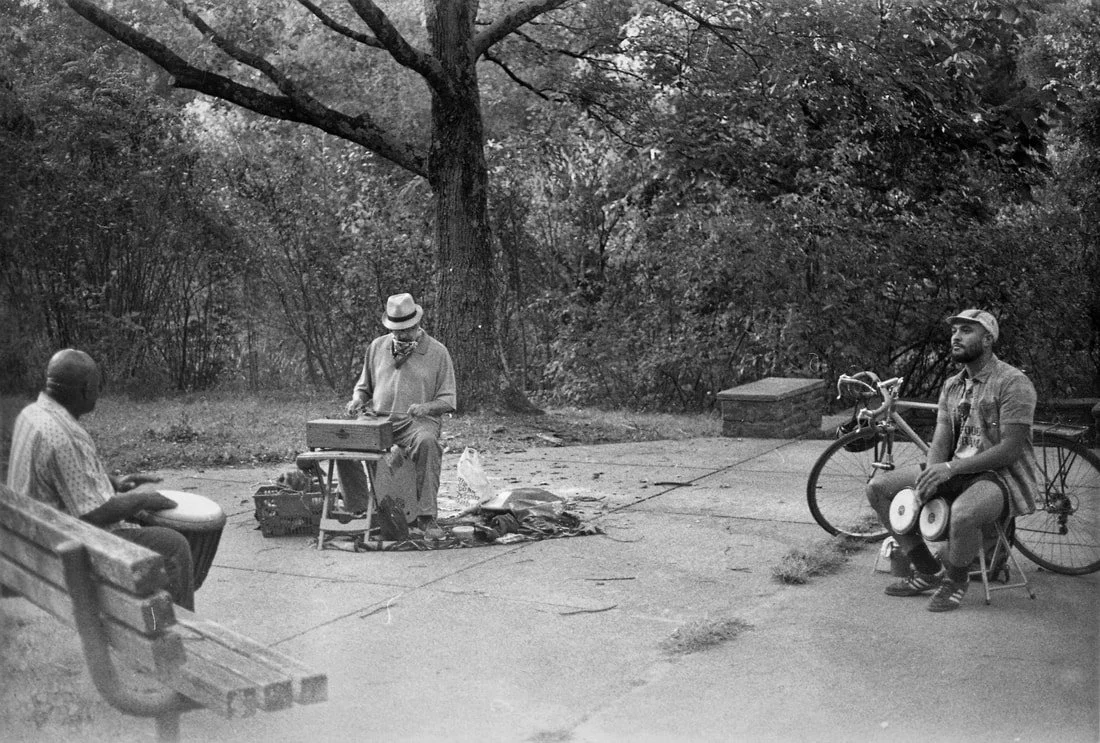 Three men are in a park, two sitting on benches and one playing a drum. The third man is sitting on the ground next to a bicycle, with a large tree in the background.