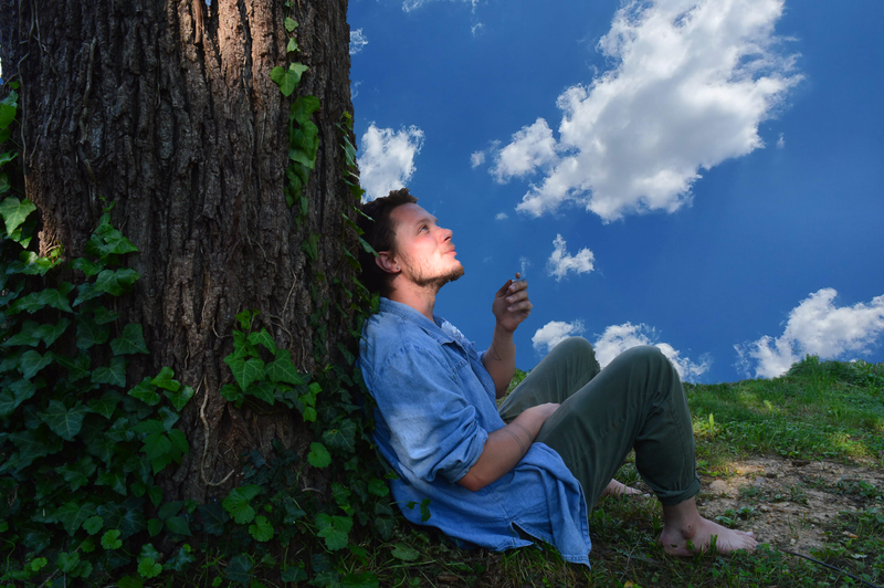 A woman with curly hair and wearing a blue shirt and green pants sitting on the ground, leaning against a large tree with ivy, looking up at the sky with clouds.