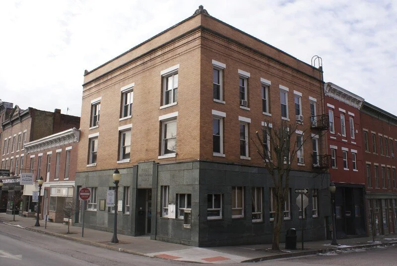 A corner building on a city street with multiple windows, trees, street lamps, and a fire escape, under a cloudy sky.