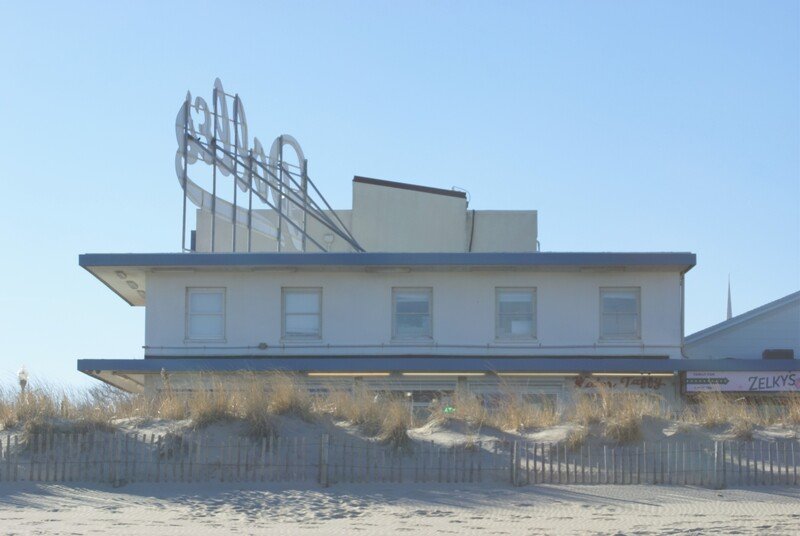 A beach house with a large neon sign on the roof that reads 'Gulf' and a sandy dune with grass in front.