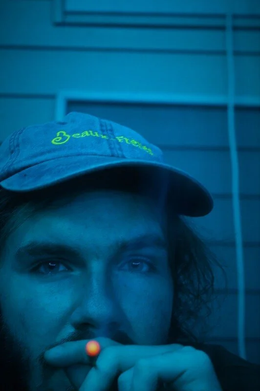 A young man wearing a baseball cap with a green embroidered logo, holding a lit cigarette, with blue lighting and a metal grid in the background.