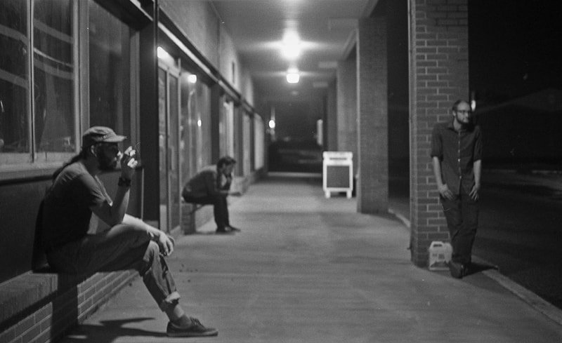 Three men waiting outside a building at night, one sitting on a ledge drinking from a bottle, one crouched down, and one standing near a brick wall.