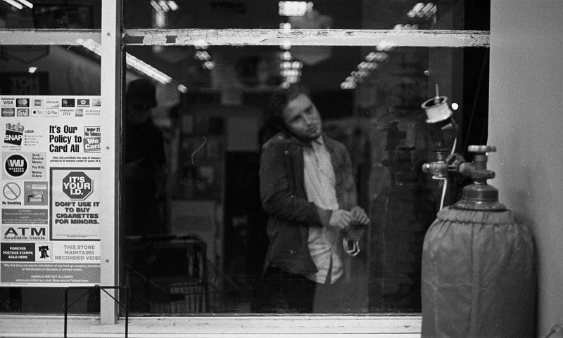 A man looking through a store window at a gas cylinder outside the store, with posters and signs on the window, reflecting the store's interior.