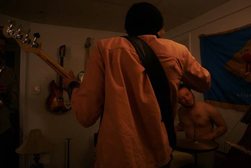 Person with black hair and tan jacket playing guitar in a dimly lit room, with a shirtless man sitting on a drum, a flag on the wall, and a wall-mounted guitar in the background.