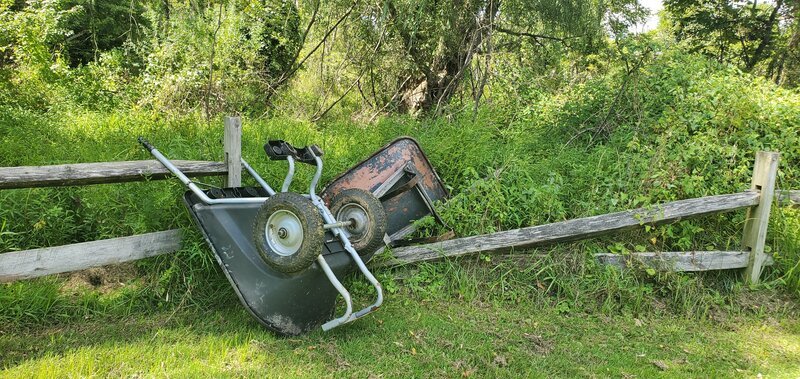 A rusty wheelbarrow resting on a broken wooden fence in a grassy area with trees in the background.