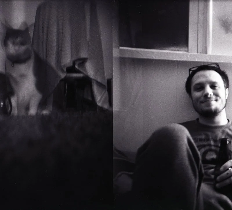 Black and white photo of a woman and a man sitting indoors, with a cat sitting beside the woman near curtains on the left, and the man smiling while holding an object on the right.