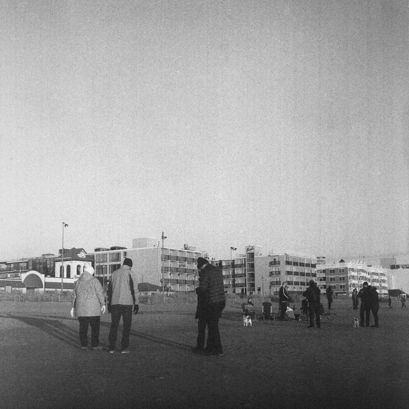 People walking and standing on a beach with modern apartment buildings in the background, black-and-white photo.