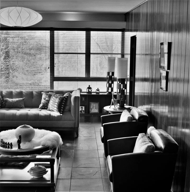 Living room with a large window, sofa with patterned pillows, armchairs, table with decorative items, lamps, and framed photographs on a shelf.
