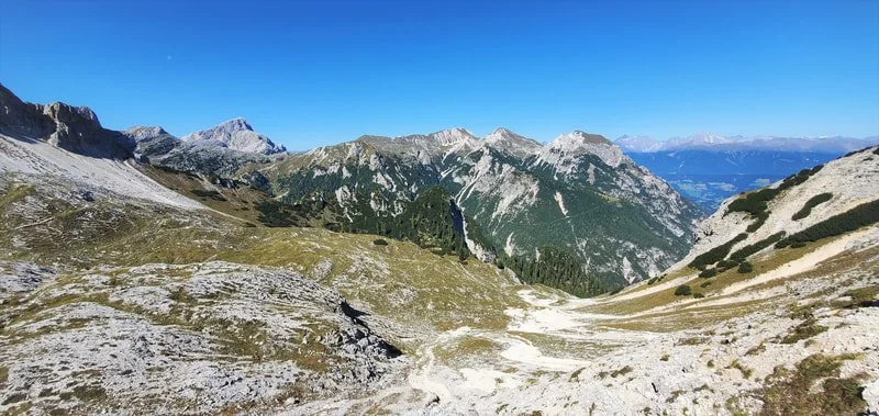 A mountain landscape with rocky and grassy terrain, forested slopes, and a clear blue sky.