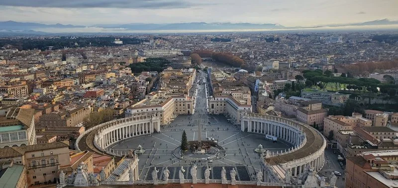 Aerial view of St. Peter's Square in Vatican City, showing the large elliptical plaza with colonnades and St. Peter's Basilica in the background.
