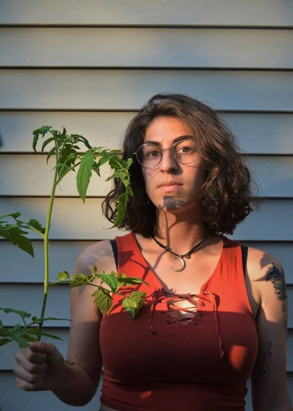 A young woman with glasses and curly brown hair standing outside against a cream-colored house with horizontal siding, holding a small green plant with her left hand, wearing a red sleeveless top, a black choker with a crescent moon pendant, and has 