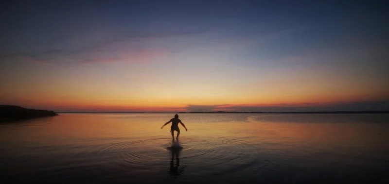 Person wading through shallow water at sunset or sunrise, with a colorful sky and calm water.