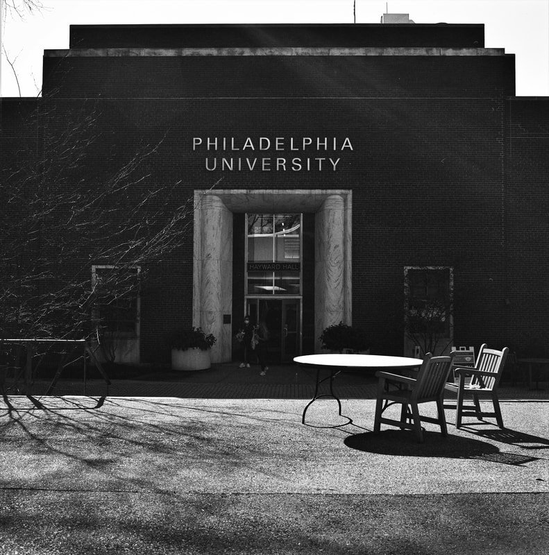 Black and white photo of the entrance to Philadelphia University with a sign above the door, outdoor chairs and tables, and shadows on the ground.