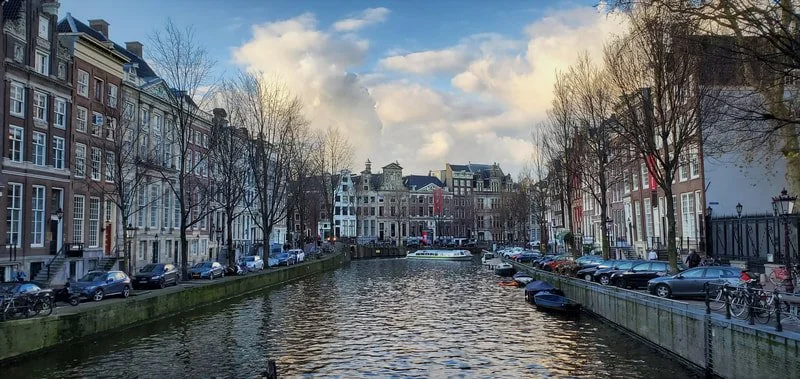 A canal lined with cars and bicycles, with historic buildings and leafless trees on either side, under a partly cloudy sky.
