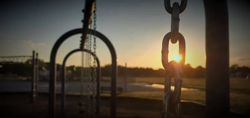 Close-up of metal playground swings and chains at sunset with a blurred background of open field and sky.