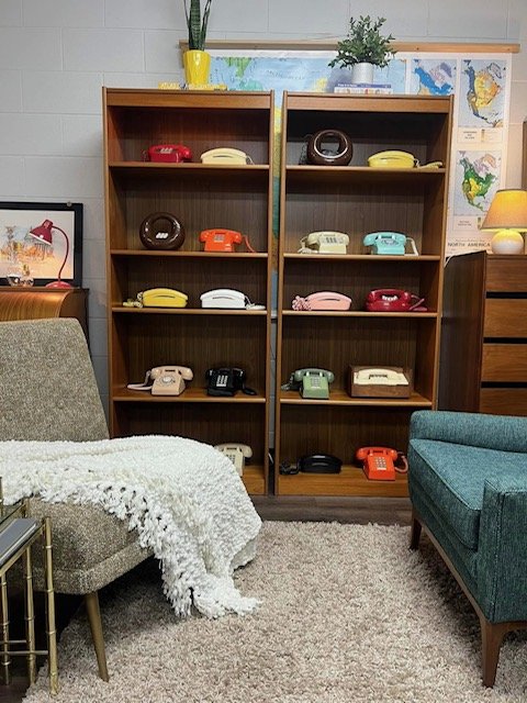 Two wooden bookshelves filled with vintage rotary phones in various colors, surrounded by mid-century modern furniture and a world map on the wall.