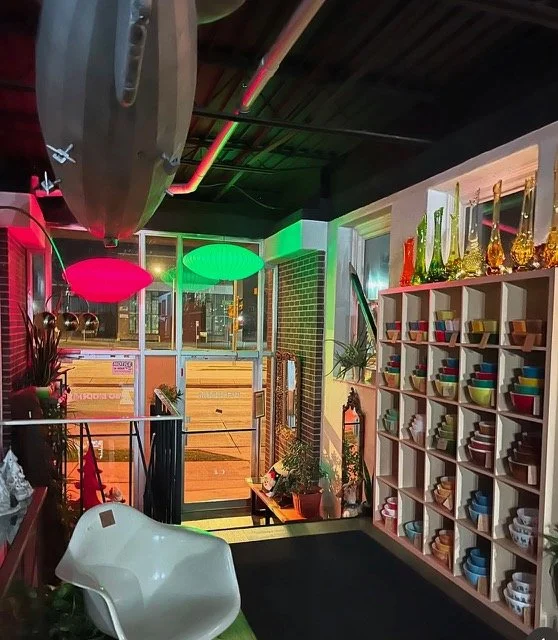 Colorful restaurant interior with neon pink and green lights, shelves with various ceramic bowls, potted plants, and a surfboard hanging from the ceiling.
