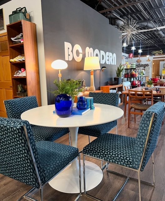 A round white table with four blue patterned chairs, a blue vase with green plants, and blue decorative items in a furniture store or showroom.