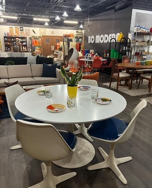 A round white table with a yellow vase holding a green plant, four glass cups, and four colorful plates. Four white and blue chairs are around the table. In the background are a sofa, various furniture pieces, and a sign that says 'BC MODERN' in a fu