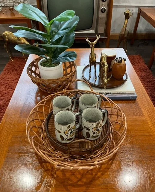 A wooden table holding a potted plant, decorative animal figures, a bowl with black pins, a rectangular tray, a stack of books, and a wicker basket with four ceramic mugs.