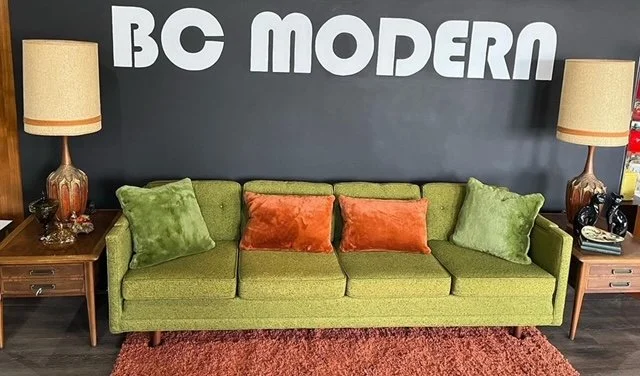 A vintage green sofa with three seat cushions and four throw pillows, flanked by matching wood side tables with lamps, in front of a dark wall with large white letters spelling 'BC MODERN'.