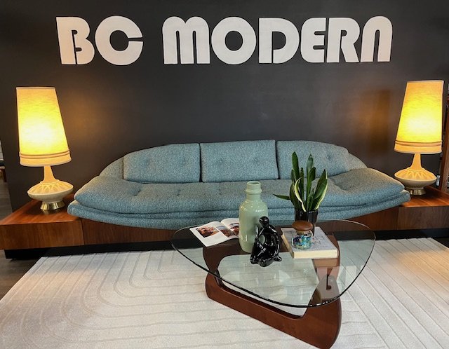 Living room with a restored mid century modern Adrian Pearsall couch, two yellow table lamps, a glass coffee table with a potted plant, a white vase, a book, and decorative items, and a hand painted mural wall sign reading 'BC MODERN'.
