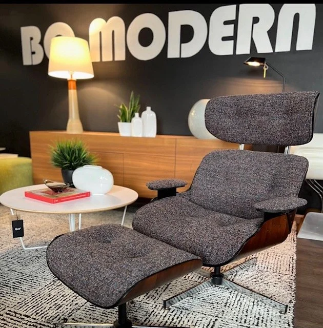Modern living room with a lounge chair, side table with a book and plant, wooden furniture with decorative vases, lamp, and wall art saying 'BOOM MODERN'.