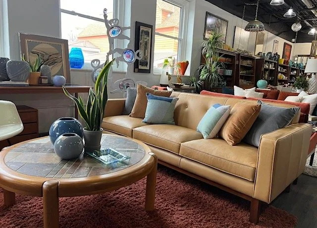 Living room with a beige sofa, decorative pillows, a round wooden coffee table with vases and a plant, and large windows in the background.