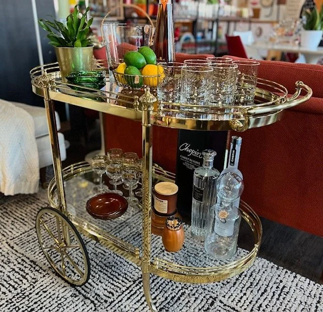 A gold bar cart with two shelves holding glassware, a potted plant, lemons, limes, and various bottles, situated in a living room with a patterned rug and red sofa.