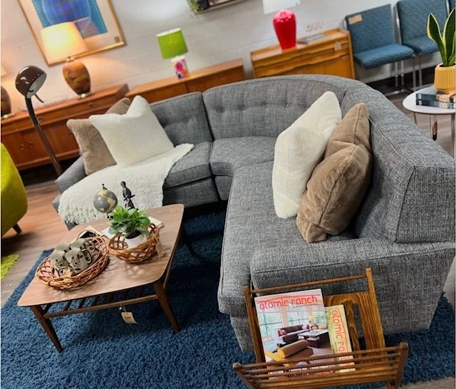 Living room with a large gray sectional sofa, wooden coffee table with decorative trays and plants, colorful lamps on wooden sideboards, and framed art on the wall.