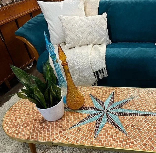 Decorative living room interior featuring a coffee table with mosaic pattern, a potted snake plant, and two colorful glass vases. A blue sofa with white pillows and throw blanket is in the background.