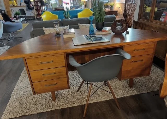 A wooden desk with three drawers on each side, placed on a beige rug. There is a gray chair in front of the desk, with a book, closed notebook, and other small items on top. In the background, there are colorful chairs, a bookshelf, plants, and a lam