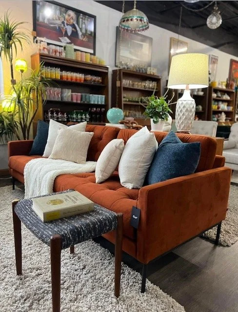 A cozy living room setting with a rust-colored sofa adorned with various pillows and a blanket, a side table with a book, a table lamp, and a potted plant, with shelves filled with colorful glassware and decor in the background.