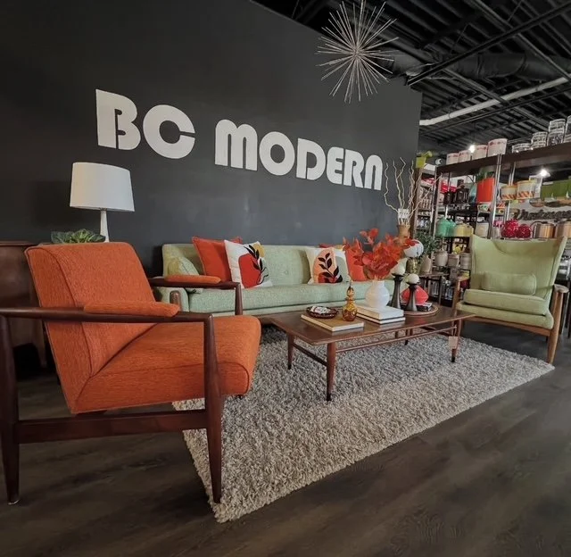 Living room setup with an orange armchair, a light green sofa with decorative pillows, and a light green armchair, all arranged around a wooden coffee table with vases and decorative items on a beige textured rug. A dark wall behind features large wh