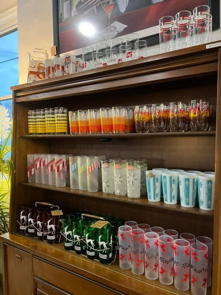 A wooden shelf with Christmas-themed glasses arranged in rows, featuring red, white, and green designs including reindeer, snowflakes, and candy canes, in a store or cafe setting.