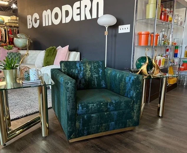 A modern living space with a dark gray wall that has bold white letters reading 'BC MODERN'. There is a teal velvet armchair with gold legs, a white sofa with colorful pillows, a glass side table with a potted plant and decorative items, a tall white