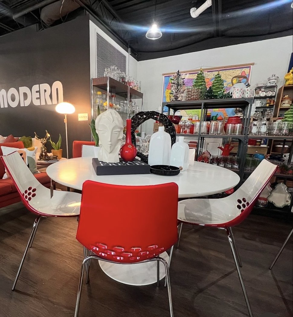 Modern dining room with a white round table, red and white chairs, and decorative items including a white sculpture of a face, vases, and glassware. Shelves and a map are visible in the background.