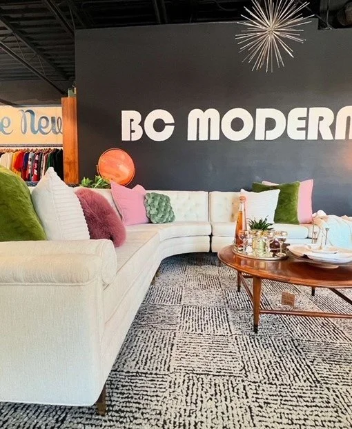 A modern lounge area with a white sectional sofa decorated with colorful pillows, a round wooden coffee table with drinks and decor, and a dark wall with 'BC MODERN' in large white letters. There is a black and white patterned rug and decorative ligh