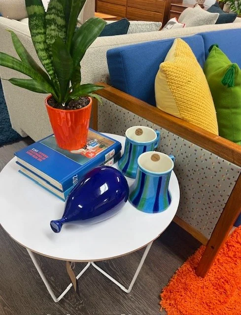 A white round table with a potted green plant, two books, two striped cups with lids, and a blue glass bottle-shaped object. In the background, a sofa with colorful pillows and a wooden side table are visible.