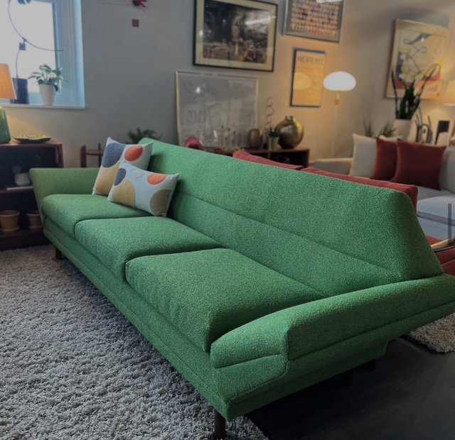 A vintage green sofa with a curved back and three cushions, in a cozy living room with framed artwork, a wooden side table, and a lamp with round shade.