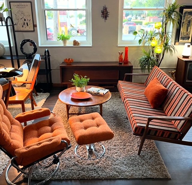 Cozy living room with vintage furniture, potted plants, and decorative items, illuminated by natural light from front windows.