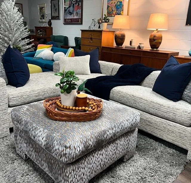 Living room with a large L-shaped gray sofa, dark blue pillows, and a matching ottoman with a woven pattern. A tray with a plant, a candle, and beads sits on the ottoman. In the background, there are colorful cushions, a white Christmas tree, and woo