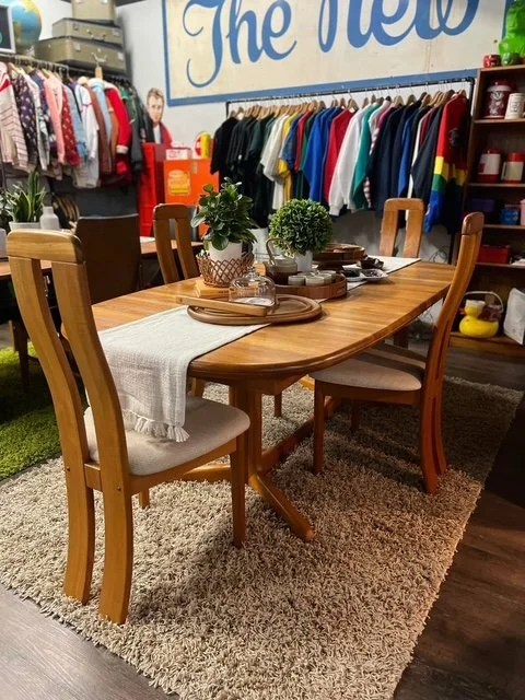 Wood dining table with four chairs, decorated with potted plants, tray, and tableware, set on a shaggy rug inside a boutique clothing store featuring hanging garments and shelving.