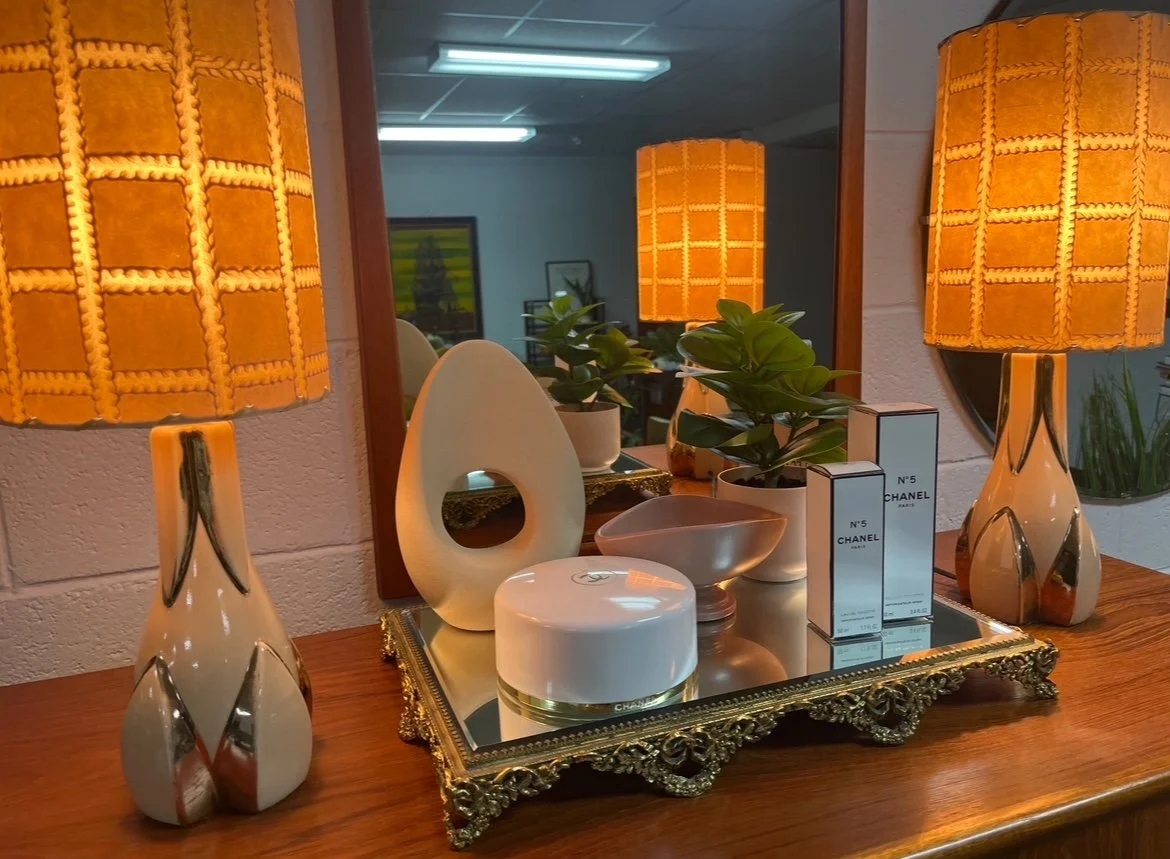 Decorative setup on a wooden table with two matching orange table lamps, a mirror, two potted green plants, several Chanel perfume boxes, a white jar, and ceramic bowls, all arranged on a ornate gold-trimmed tray, with a wall mirror reflecting part o
