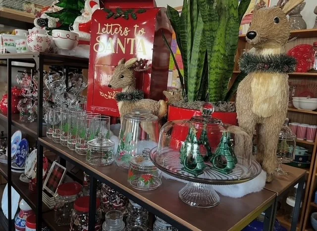 Decorative holiday display featuring two stuffed animals dressed as reindeer with Christmas wreath neck accessories, a red 'Letters to Santa' mailbox, a green plant, various glass jars, and Christmas-themed ornaments and decorations on shelves.