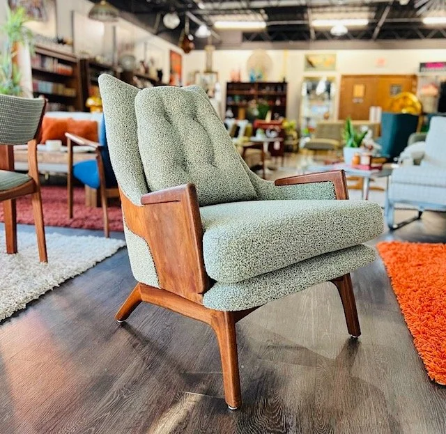 A vintage mid-century modern armchair with green upholstery and wooden armrests, in a furniture store with various chairs, sofas, and rugs in the background.