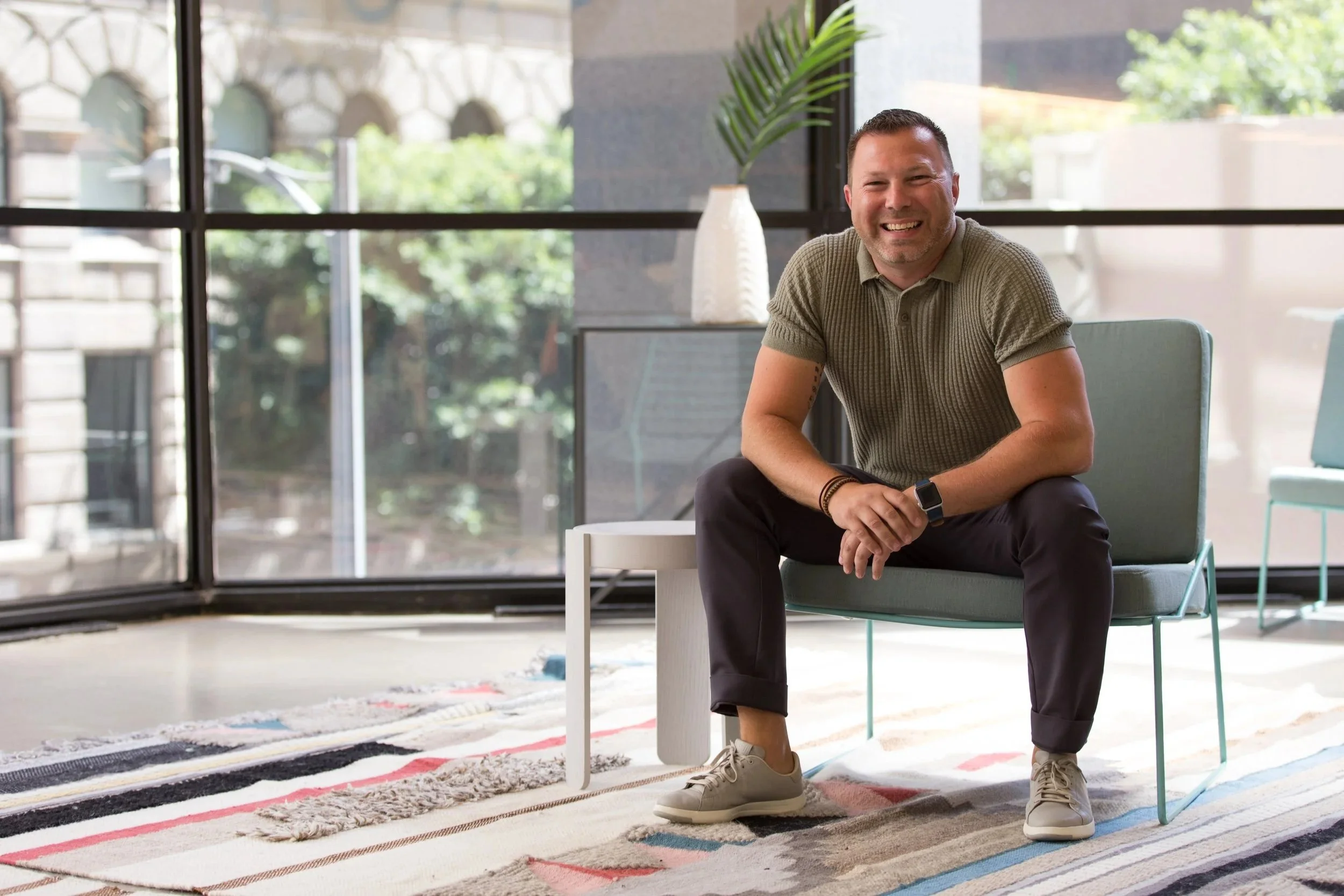 Smiling man sitting on chair near large window in modern office space, with greenery and floral rug visible inside.