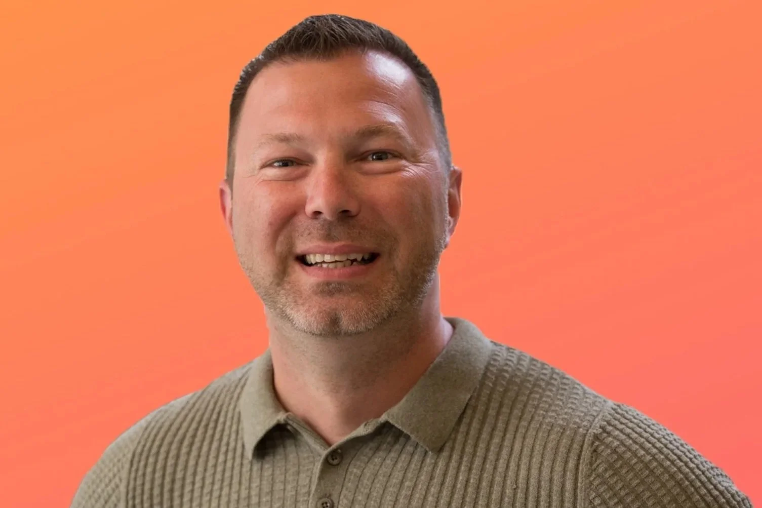 A smiling man with short brown hair and facial hair, wearing a beige textured polo shirt, against an orange background.