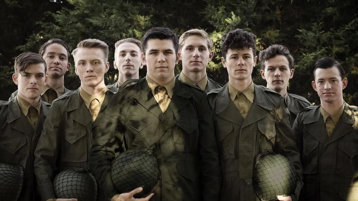 Group of nine young men in military uniforms standing outdoors with trees in the background, some holding helmets.